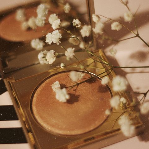 Un surligneur doré et compact avec de petites fleurs blanches sur le dessus, se reflétant dans un miroir, créant une esthétique douce et élégante.