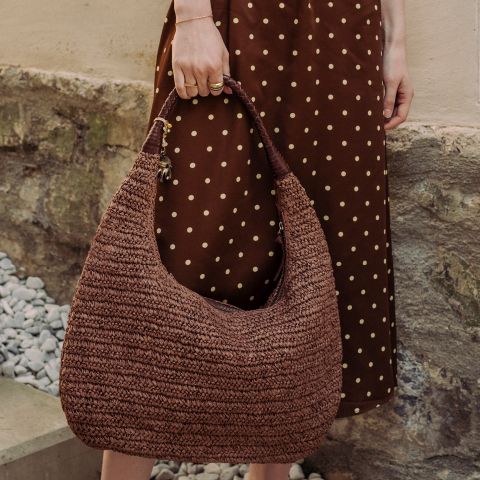 Une personne en robe marron à pois tient un grand sac à main tissé marron, tandis qu'elle se tient à côté d'un mur de pierre et de gravier.