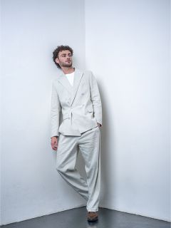 Un homme aux cheveux bouclés, vêtu d'un costume gris clair et d'un t-shirt blanc, est appuyé contre un mur blanc dans un environnement de studio minimaliste.