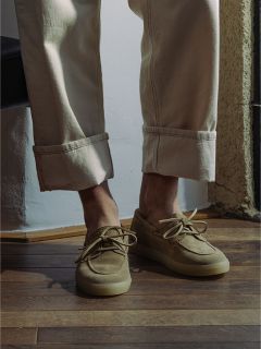 Personne portant un pantalon beige à revers et des chaussures bateau en daim marron clair, debout sur un sol en bois à proximité de la lumière du soleil.