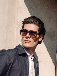 Un homme aux cheveux châtains coiffés portant de grandes lunettes de soleil carrées, une veste sombre et une chemise claire, se tenant à la lumière du soleil à côté d'un mur texturé.