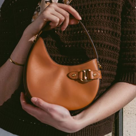 Personne avec un petit sac à main en cuir marron clair avec une fermeture à boucle, un haut en tricot marron et un bracelet doré.