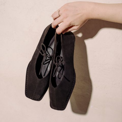 Une main tient une paire de chaussures à lacets en daim noir à bout carré devant un mur texturé beige clair.