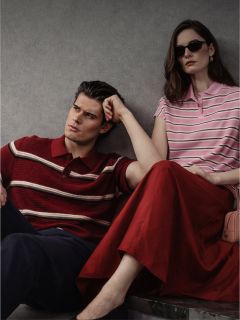 Un homme et une femme sont assis contre un mur, tous deux portant des polos rayés ; l'homme porte un pantalon foncé, la femme une jupe rouge et des lunettes de soleil.