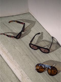 Trois paires de lunettes de soleil avec différentes montures et verres sont placées sur des marches en béton à la lumière du soleil devant un mur blanc.