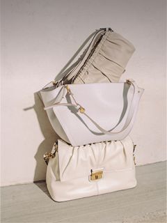 Trois sacs à main élégants de couleur crème sont empilés devant un mur clair, présentant différentes textures et des détails de quincaillerie dorée dans une lumière douce et naturelle.