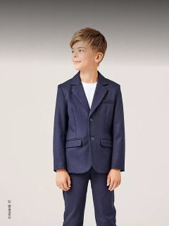 Un garçon en costume bleu marine et chemise blanche se tient là, souriant et regardant sur le côté devant un fond clair.