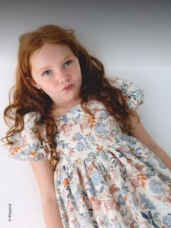 Une jeune fille aux longs cheveux roux vêtue d'une robe à fleurs se tient devant un fond blanc et fait une moue enjouée.