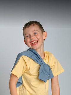 Garçon souriant en chemise jaune avec un pull bleu sur les épaules, debout devant un simple fond gris.