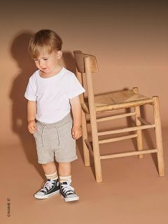 Un jeune enfant en t-shirt blanc, short gris et baskets noires se tient à côté d'une chaise en bois devant un fond beige.