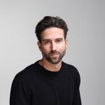 Un homme aux cheveux courts châtains et portant une barbe, vêtu d'un pull noir, est assis devant un arrière-plan gris clair uni et regarde la caméra avec une expression neutre.