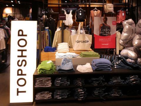 Vitrine d'un magasin de vêtements avec des hauts, des shorts et des sacs soigneusement pliés sur les étagères, un panneau lumineux "TOPSHOP" et une affiche publicitaire en arrière-plan.