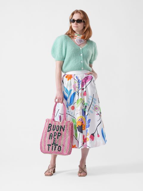 Femme portant des lunettes de soleil, un cardigan vert menthe, une jupe à fleurs, une écharpe et des sandales, et tenant un sac cabas rose avec l'inscription « BUON APPETITO ».