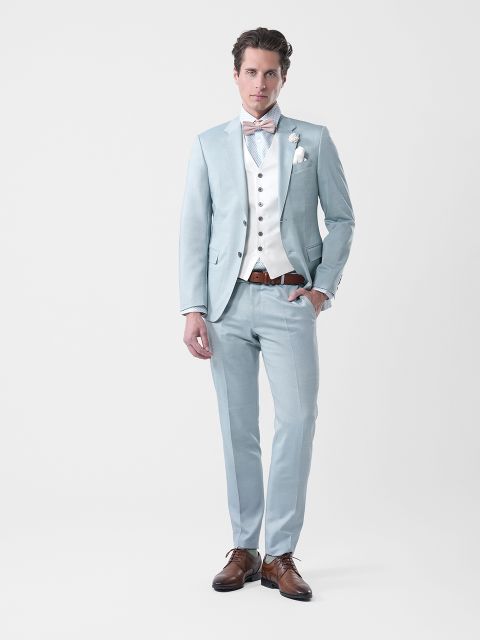 Homme en costume bleu clair, gilet blanc, nœud papillon et chaussures marron, debout devant un arrière-plan blanc sobre.