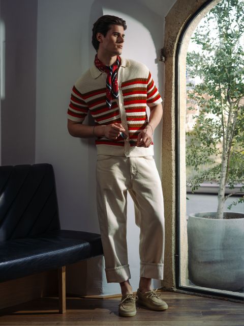 Un homme en polo tricoté rayé, pantalon crème et lunettes de soleil pose à la fenêtre avec une plante.