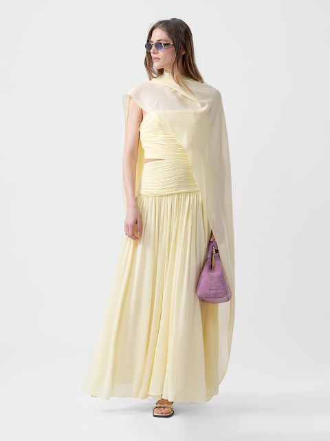 Femme vêtue d'une robe jaune pâle fluide avec une épaule drapée, des lunettes de soleil, des sandales et un petit sac à main violet devant un arrière-plan sobre.