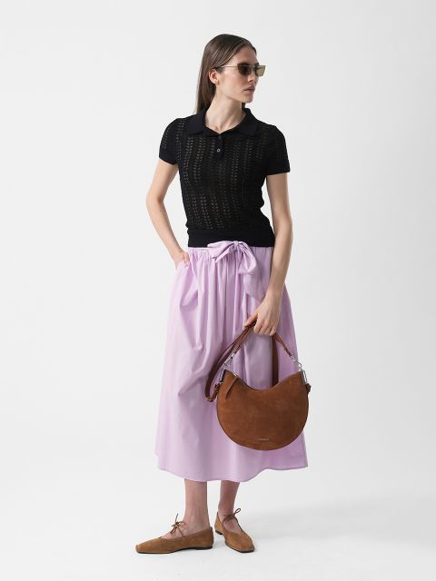 Femme portant des lunettes de soleil, un haut noir à manches courtes, une jupe violet clair et des chaussures marron, tenant un sac à main marron en forme de croissant devant un fond blanc.