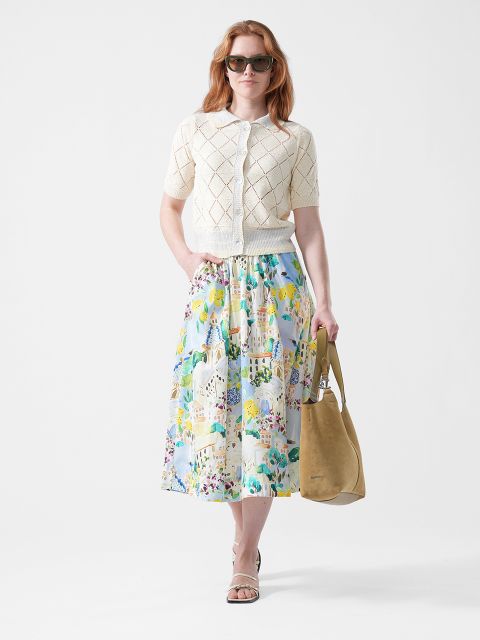 Femme portant des lunettes de soleil, un cardigan tricoté à manches courtes de couleur crème, une jupe fleurie colorée, des sandales et un sac fourre-tout marron clair sur fond blanc.