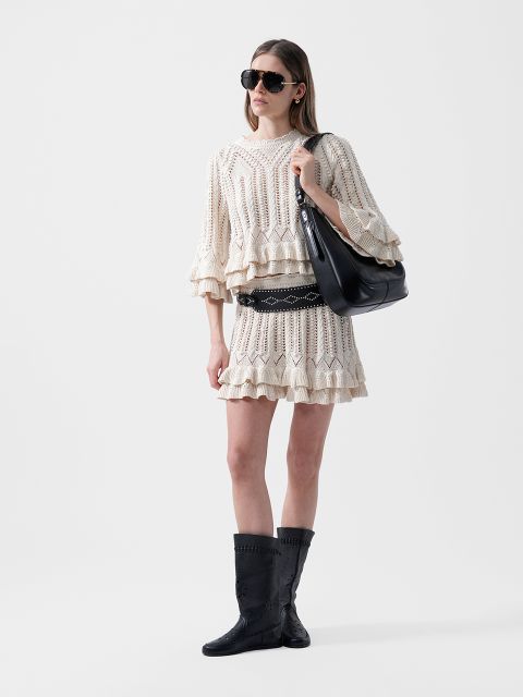 Une femme en costume de crochet crème, bottes noires, lunettes de soleil et grand sac noir se tient devant un simple fond blanc.