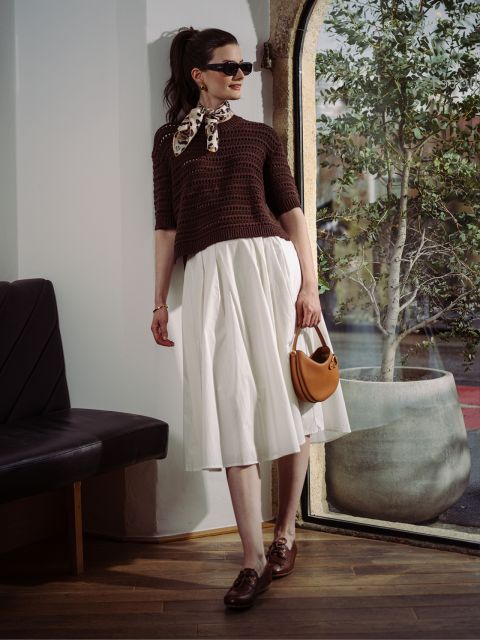Une femme avec des lunettes de soleil, un pull marron et une jupe blanche pose près d'une fenêtre avec un arbre.
