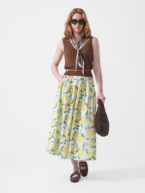 Femme portant des lunettes de soleil, un haut marron sans manches et une longue jupe à imprimé citron, tenant un sac marron à la main et portant un foulard autour du cou, debout devant un fond blanc.