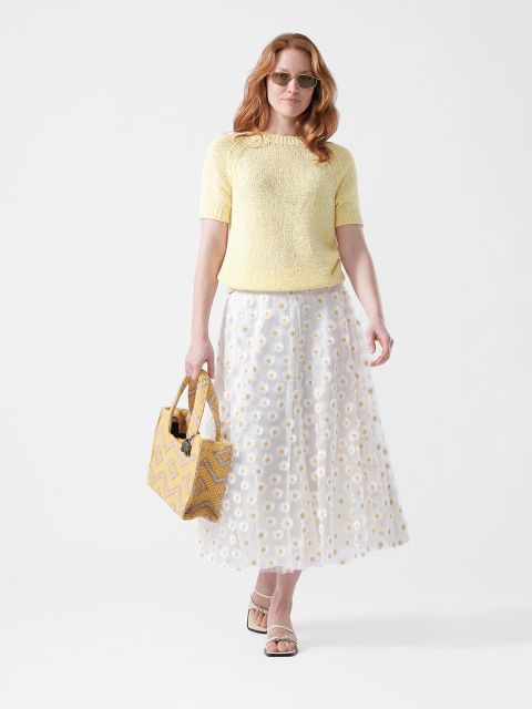 Femme avec des lunettes de soleil, portant un haut jaune, une jupe blanche à motifs de marguerites et des sandales, tenant un sac fourre-tout à motifs jaunes devant un fond blanc.