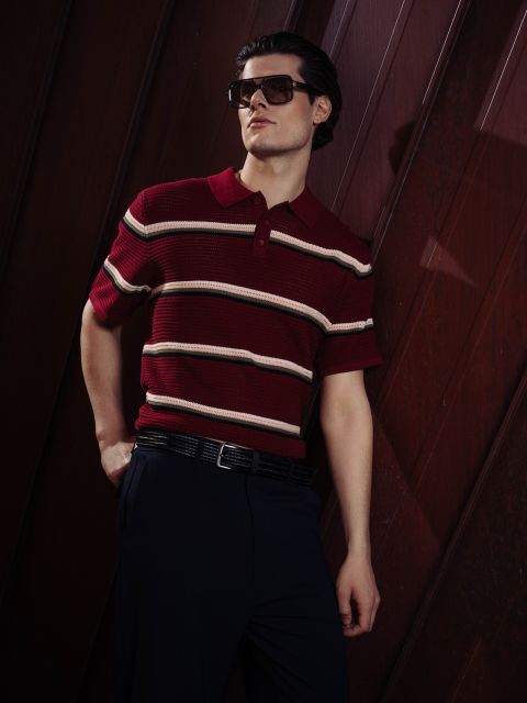 Un homme portant des lunettes de soleil et un polo à rayures rouges pose avec assurance, une main dans la poche, devant un fond sombre en lambris.