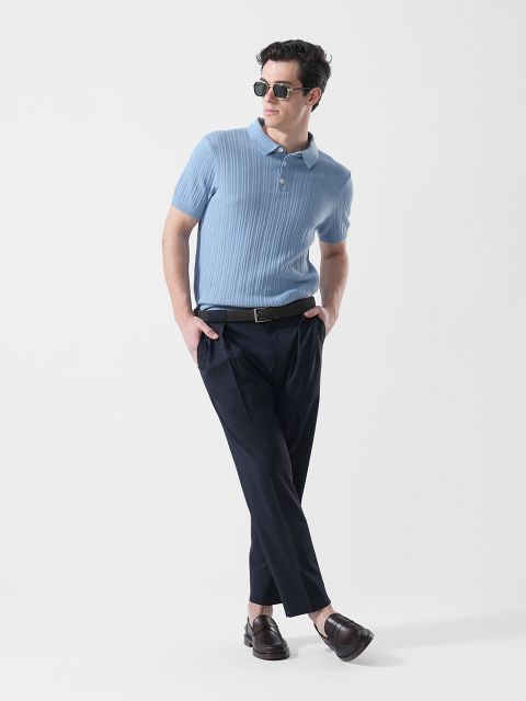 Homme avec des lunettes de soleil, portant un polo bleu clair à manches courtes, un pantalon foncé et des mocassins marron, debout avec un pied croisé devant l'autre.