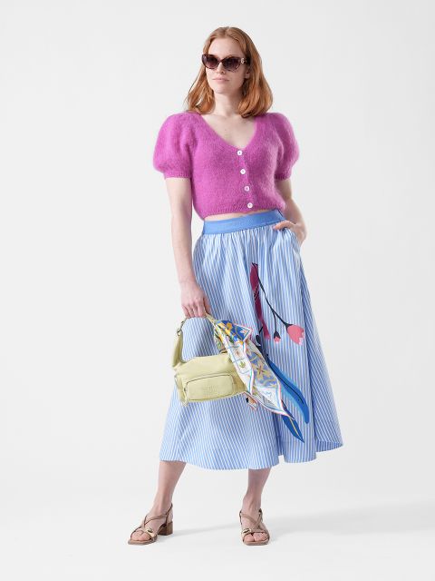 Femme portant un haut duveteux rose, une jupe midi bleue à rayures avec un motif floral, des lunettes de soleil, un sac jaune avec un foulard et des sandales.