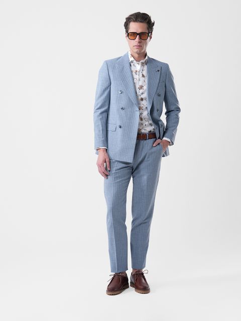 Un homme en costume bleu clair, chemise blanche à motifs, chaussures marron et lunettes de soleil, debout avec une main dans la poche devant un fond uni.