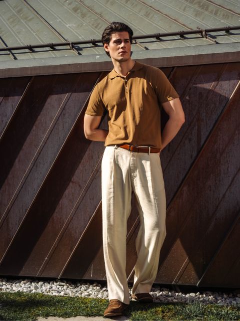 Un homme se tient à l'extérieur, vêtu d'un polo marron, d'un pantalon à pinces de couleur crème et de chaussures marron, et s'appuie contre un bâtiment métallique à la lumière du soleil.