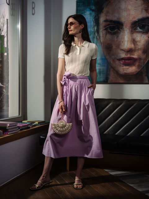 Une femme porte un polo crème et une jupe lavande fluide, tenant un sac à main en forme d'éventail orné de perles.