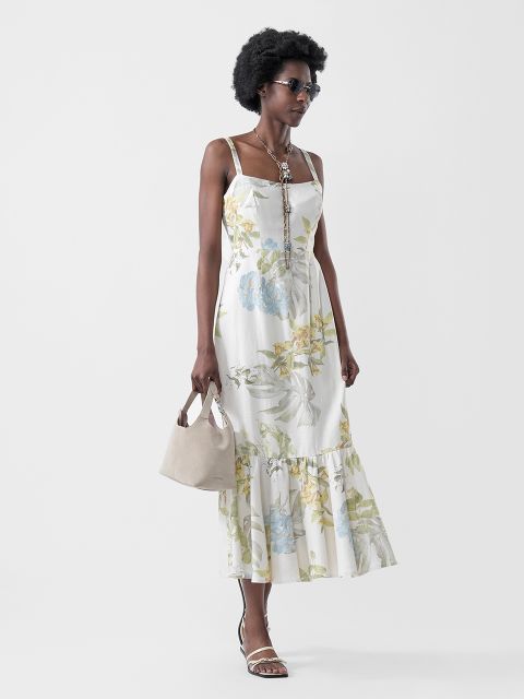Une femme en robe d'été blanche à fleurs, sandales, lunettes de soleil et collier, portant un sac à main beige, pose devant un fond clair uni.