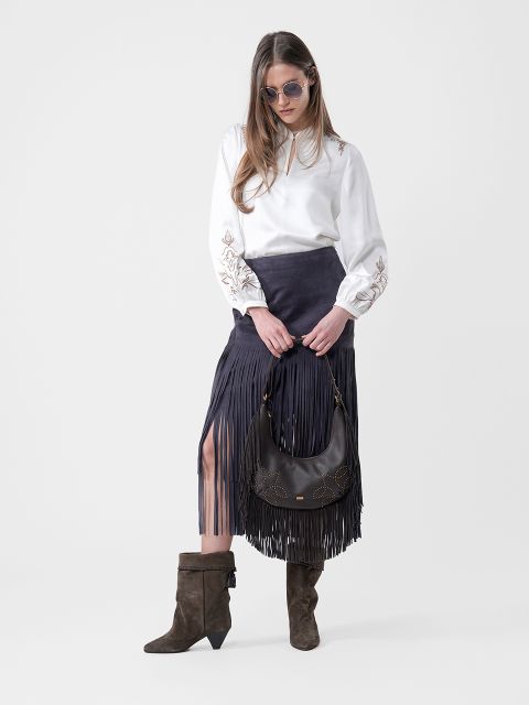 Femme portant des lunettes de soleil, un chemisier blanc, une jupe noire à franges, des bottes marron et un sac à main noir à la main, devant un arrière-plan sobre.