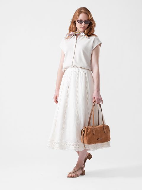 Une femme portant des lunettes de soleil, un chemisier blanc et une jupe longue blanche, tenant un sac à main marron clair et portant des sandales beiges, se tient devant un fond blanc.