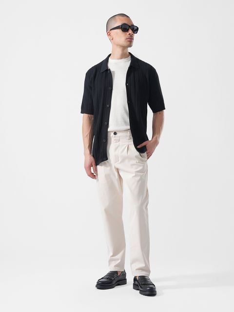 Homme portant des lunettes de soleil, une chemise noire à manches courtes sur un t-shirt blanc, un pantalon couleur crème et des chaussures noires, debout devant un fond blanc.