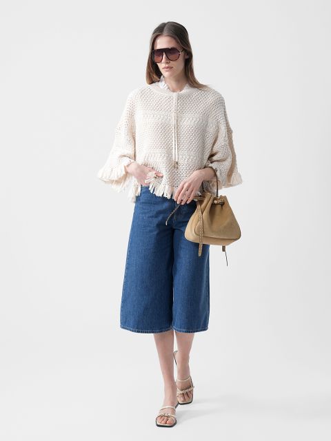 Une femme porte des lunettes de soleil, un pull en tricot crème à franges, un jean-culotte bleu longueur genou et des sandales, et tient un sac à main marron clair devant un fond blanc.