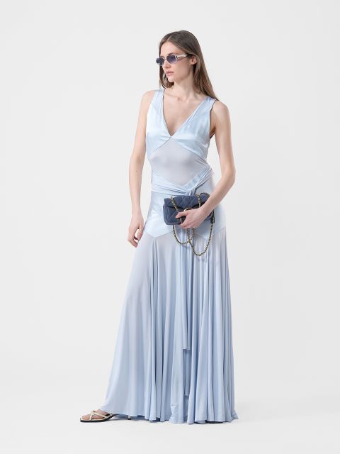 Femme portant une robe bleu clair fluide sans manches avec des lunettes de soleil, tenant un sac à main foncé avec une chaîne, debout devant un fond blanc uni.