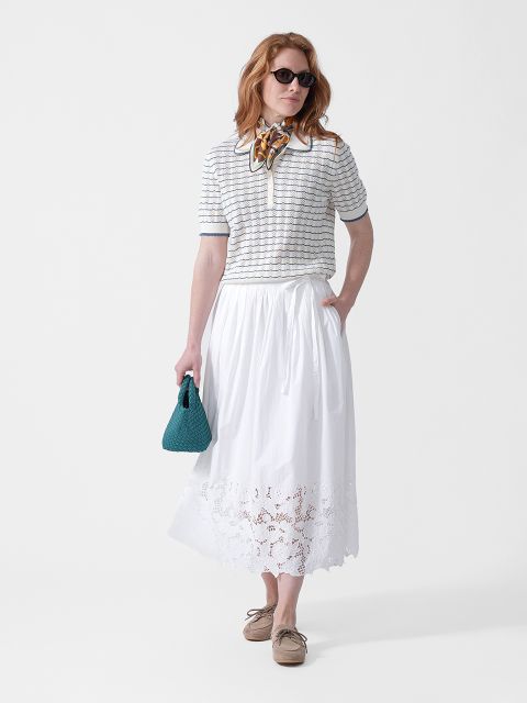 Une femme porte des lunettes de soleil, un haut rayé, une jupe midi blanche brodée, un foulard autour du cou, un sac sarcelle et des chaussures beiges.