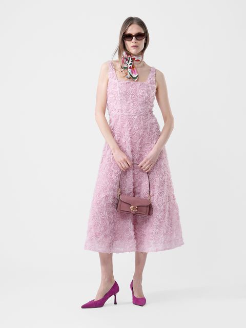 Femme dans une robe rose texturée sans manches, des talons violets, des lunettes de soleil, une écharpe autour du cou et tenant un sac à main violet, debout devant un fond blanc.