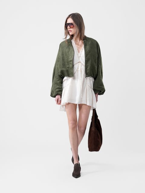 Femme vêtue d'une robe blanche, d'une veste verte, de lunettes de soleil et de bottes marron, portant un grand sac marron devant un arrière-plan blanc uni.