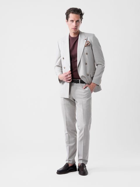 Un homme en costume gris clair et chemise marron pose avec assurance devant un fond blanc, une main sur sa veste, l'autre dans sa poche.