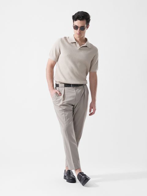 Un homme portant des lunettes de soleil, une chemise en maille beige à manches courtes, un pantalon gris clair et des mocassins noirs se tient devant un simple fond blanc.