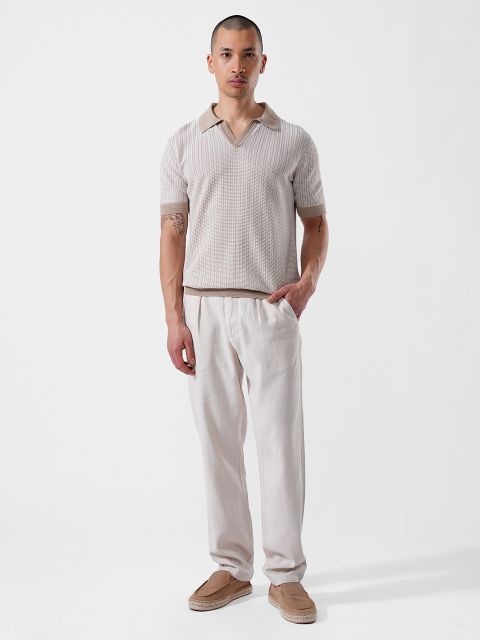 Un homme se tient devant un fond blanc et porte une chemise beige à manches courtes avec un col, un pantalon clair et des mocassins marron clair.