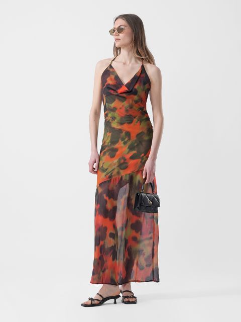 Une femme vêtue d'une maxi-robe sans manches à motifs orange et verts, de sandales et de lunettes de soleil, tenant un petit sac à main noir, se tient devant un fond blanc.