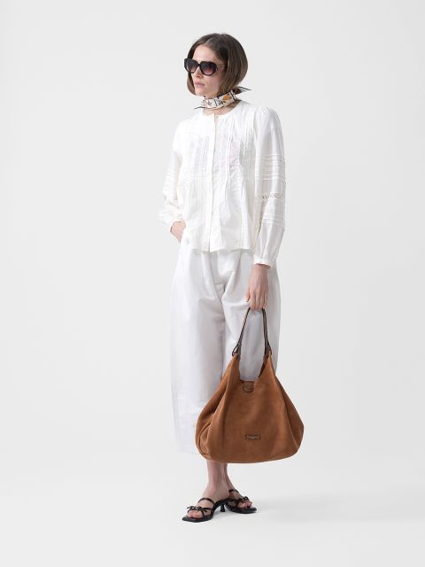 Femme portant des lunettes de soleil, un chemisier blanc et un pantalon, une écharpe autour du cou, des sandales noires et tenant à la main un grand sac à main en daim marron.