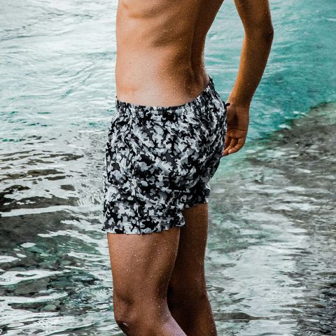 Une personne se tenant dans l'eau peu profonde et portant un maillot de bain camouflage noir et blanc, son torse n'étant pas visible sur l'image.