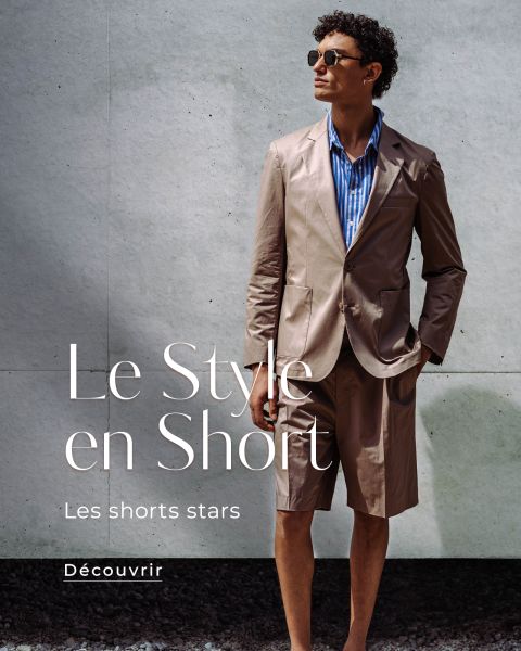 Un homme en costume marron clair avec un short, une chemise rayée bleue et des lunettes de soleil se tient devant un mur gris. Texte : "Le Style en Short, Les shorts stars."