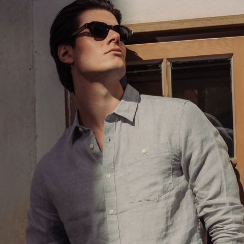 Un homme avec des lunettes de soleil et une chemise boutonnée gris clair se tient à la fenêtre et regarde le soleil.