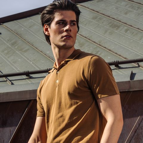 Un homme dans un polo marron à manches courtes se tient dehors devant un toit en métal et regarde de côté avec un air sérieux.